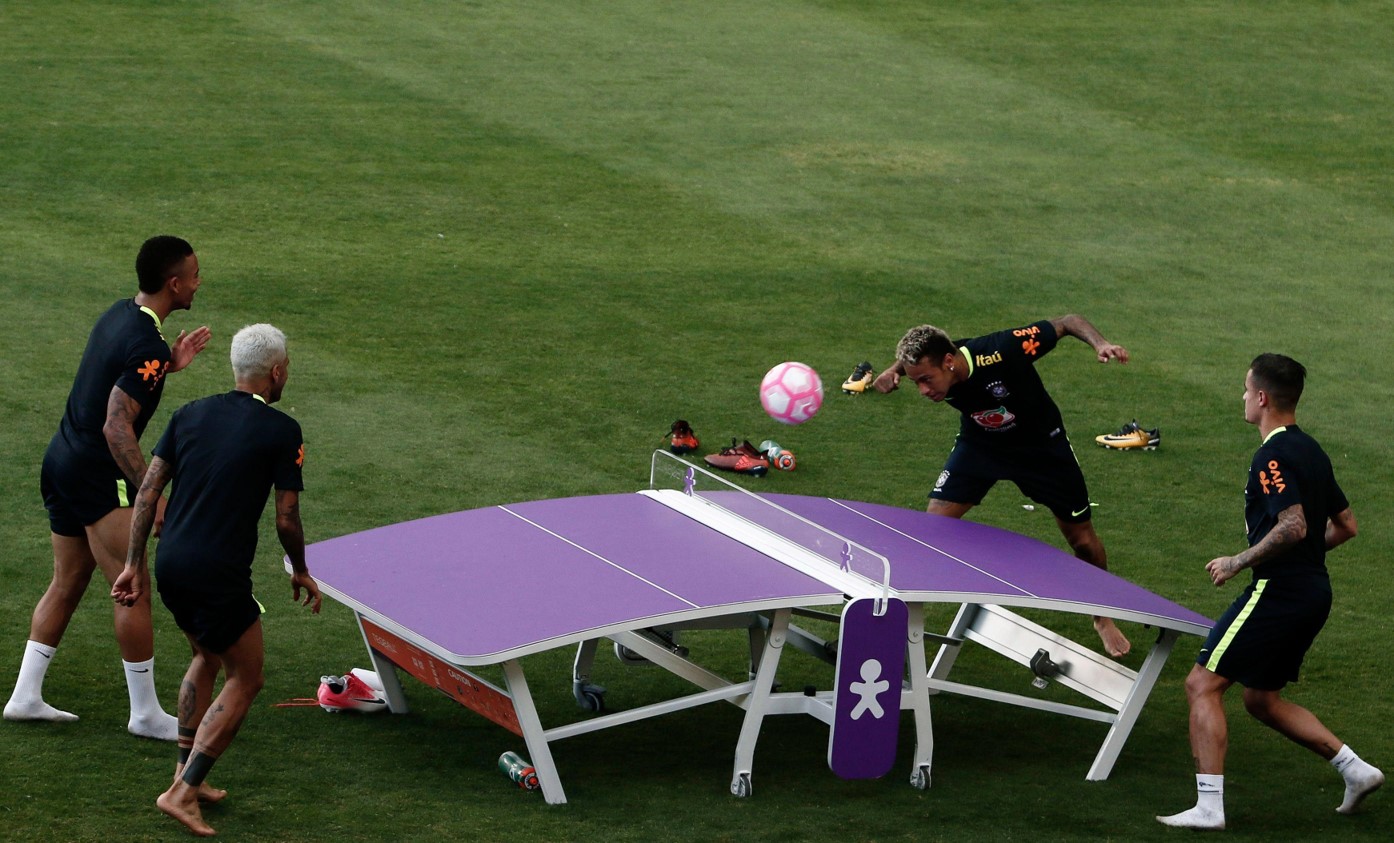 Neymar bate na bola com a cabeça em uma mesa semelhante a de ping pong com dois atletas da seleção brasileira do lado oposto e um ao seu lado