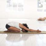 mulher em sala bastante branca faz posição de yoga sentada no chão com as mãos esticadas para frente e a cabeça para baixo