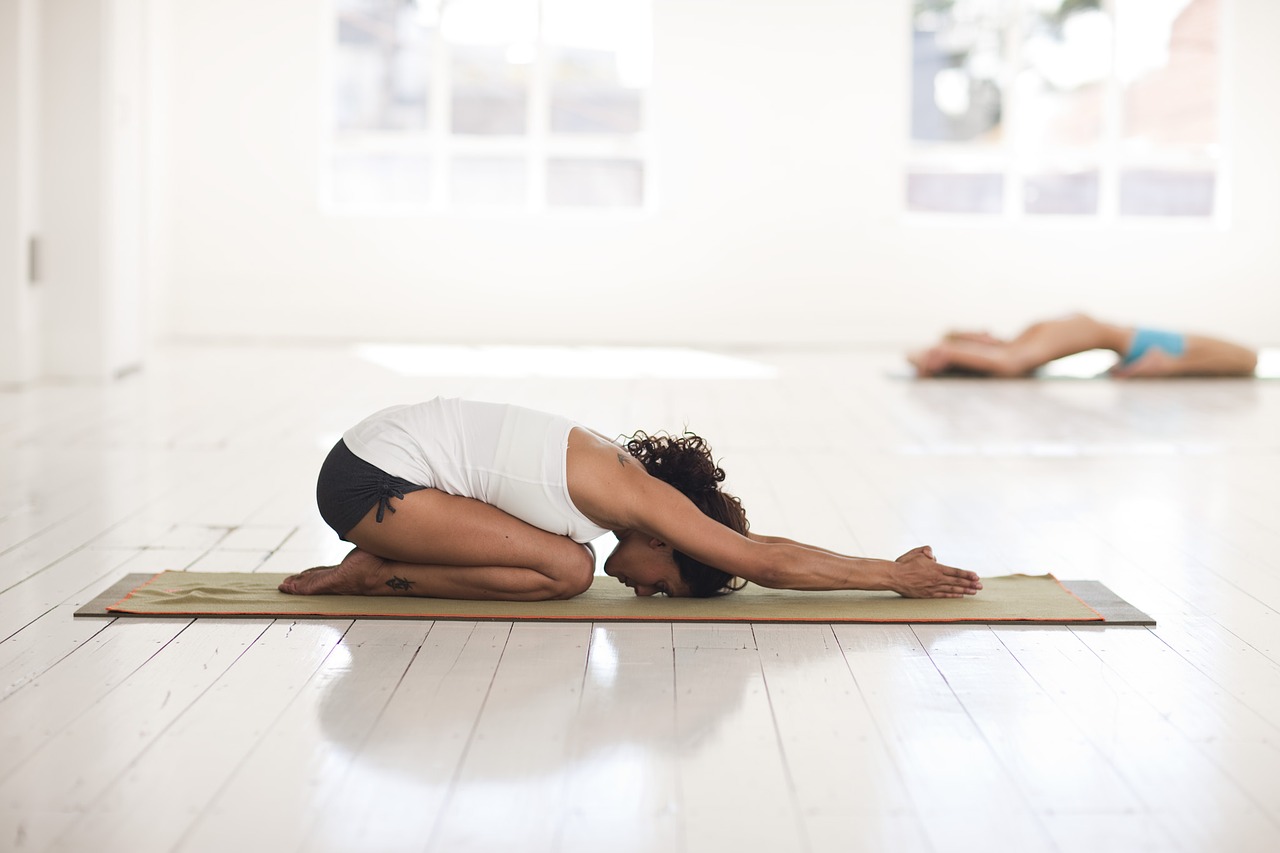 mulher em sala bastante branca faz posição de yoga sentada no chão com as mãos esticadas para frente e a cabeça para baixo
