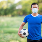 homem camisa azul e máscara segurando bola de futebol