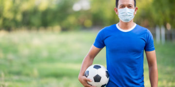 homem camisa azul e máscara segurando bola de futebol