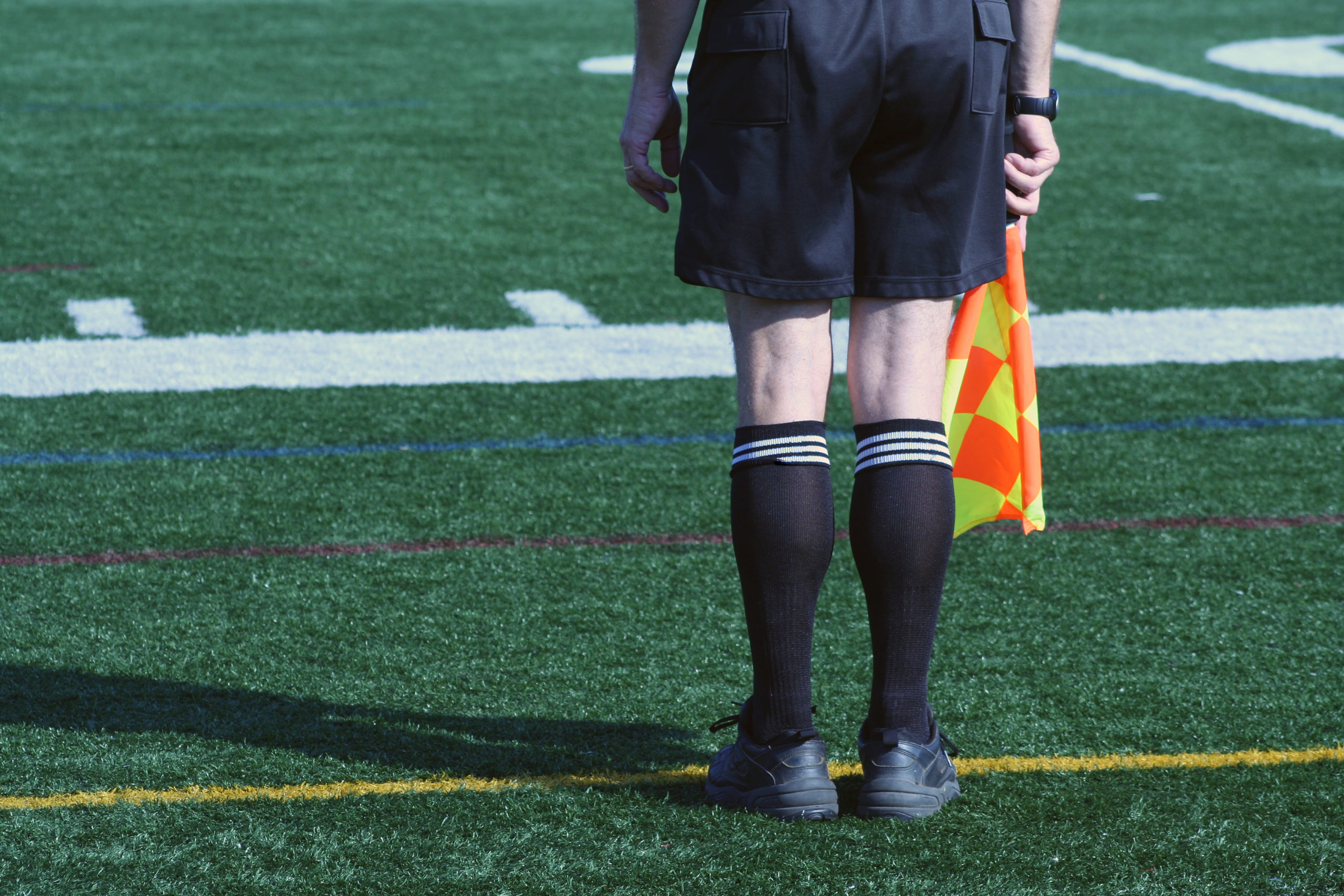 Pernas de um juíz de costas com bandeira na mão no campo de futebol