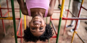 Menina negra de de roupa rosa com cerca de 5 anos de idade de cabeça para baixo brincando de se pendurar em brinquedo colorido.