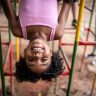 Menina negra de de roupa rosa com cerca de 5 anos de idade de cabeça para baixo brincando de se pendurar em brinquedo colorido.