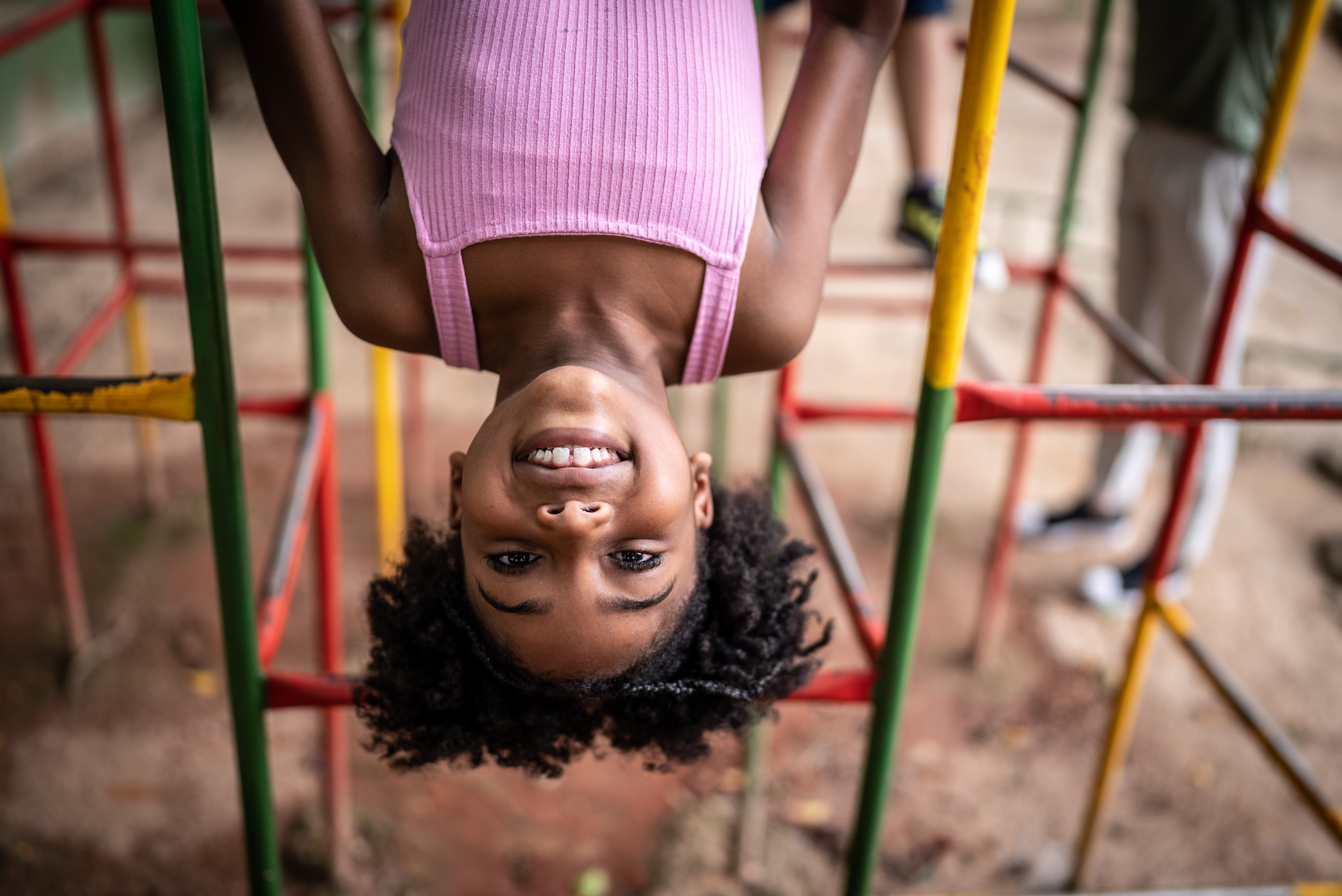 Menina negra de de roupa rosa com cerca de 5 anos de idade de cabeça para baixo brincando de se pendurar em brinquedo colorido.